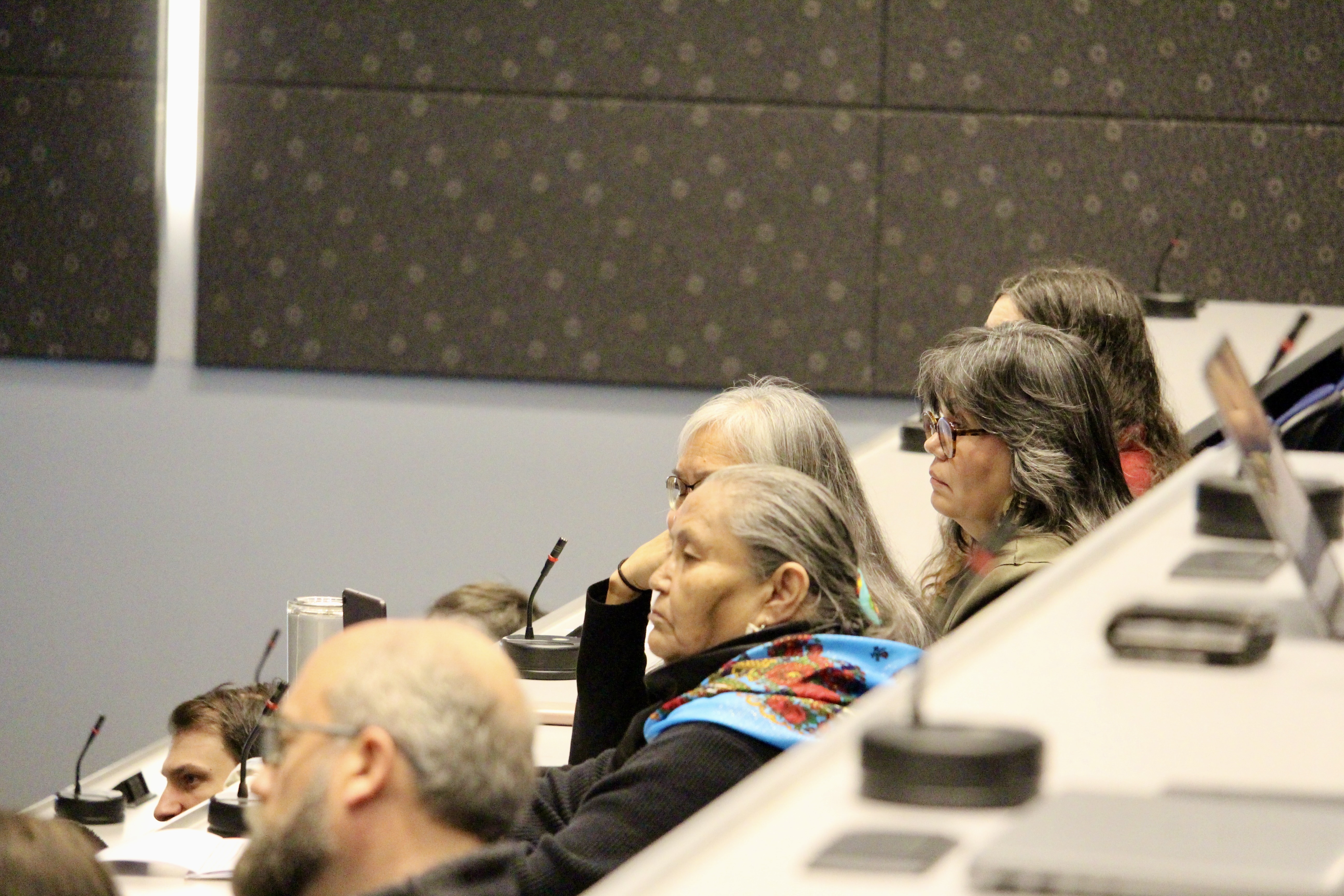 A group of people sit in the audience, including Kə̀ɬkəmpica Elder Rose Caldwell