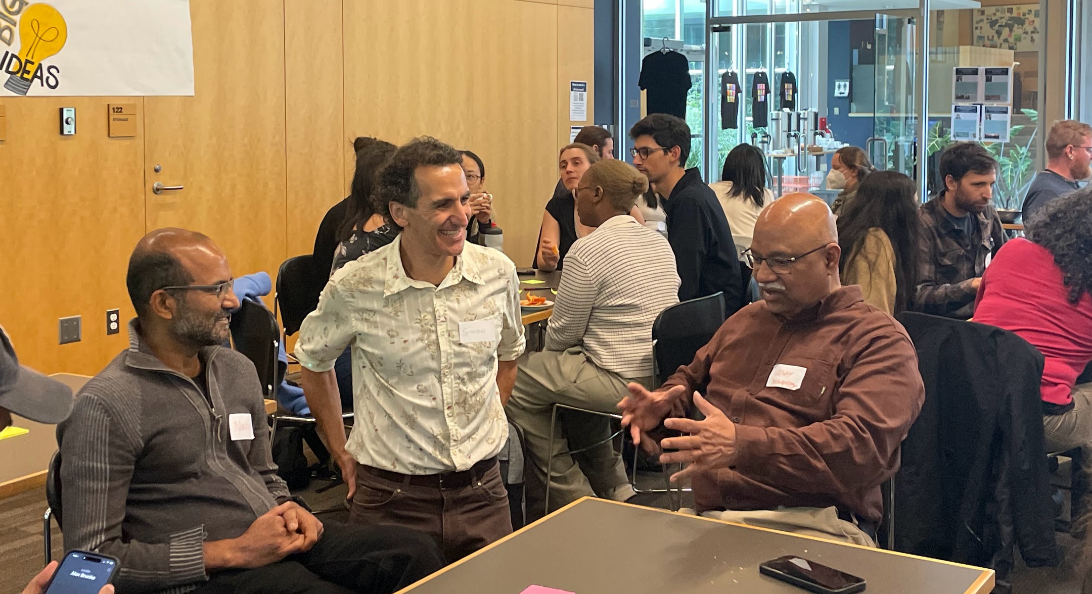 3 attendees sit around a table, with more people sitting at tables behind them in a room with wooden walls and windows. The people at the forefront are smiling and clearly mid-conversation, and are wearing name tags.