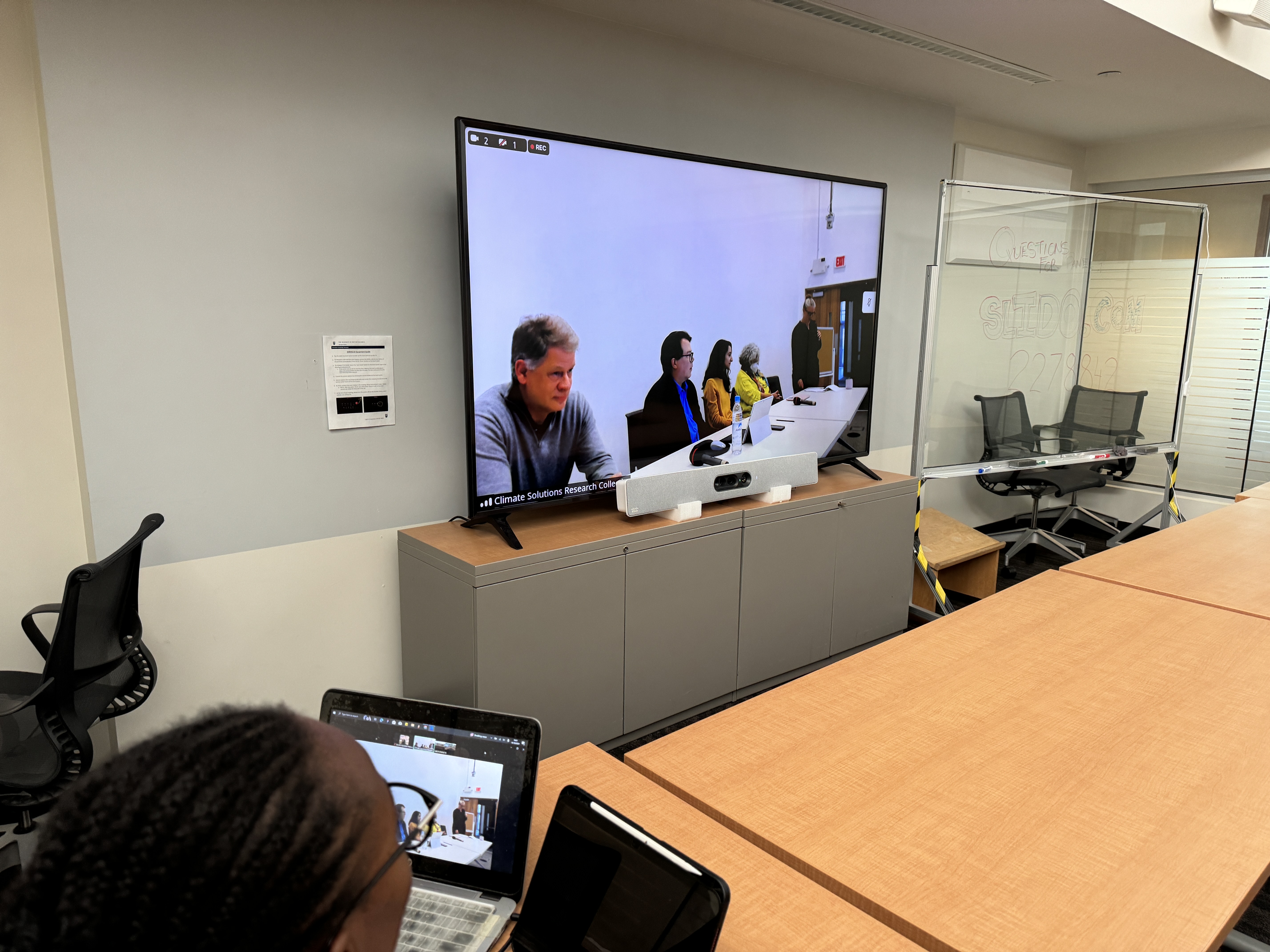 The image shows a large screen showing a panel of four speakers and one moderator. In the room there are tables, a board with marker writing on it depicting a Slido code, and one person's head.